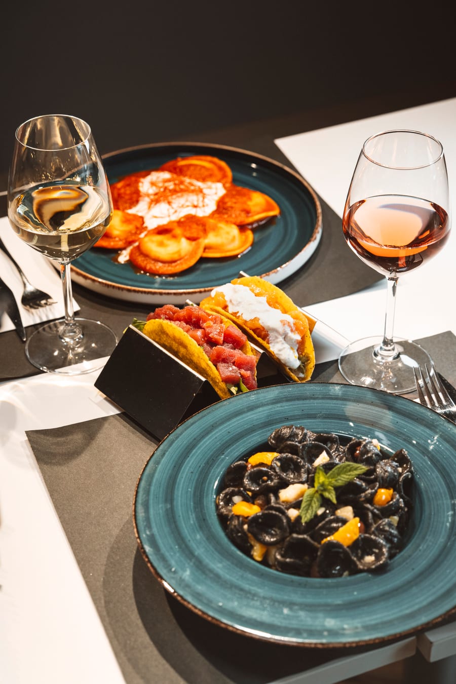 Tre piatti sulla tavola — i fritti, i taco di tartare di tonno e la pasta nera coi pomodorini gialli — fra due calici