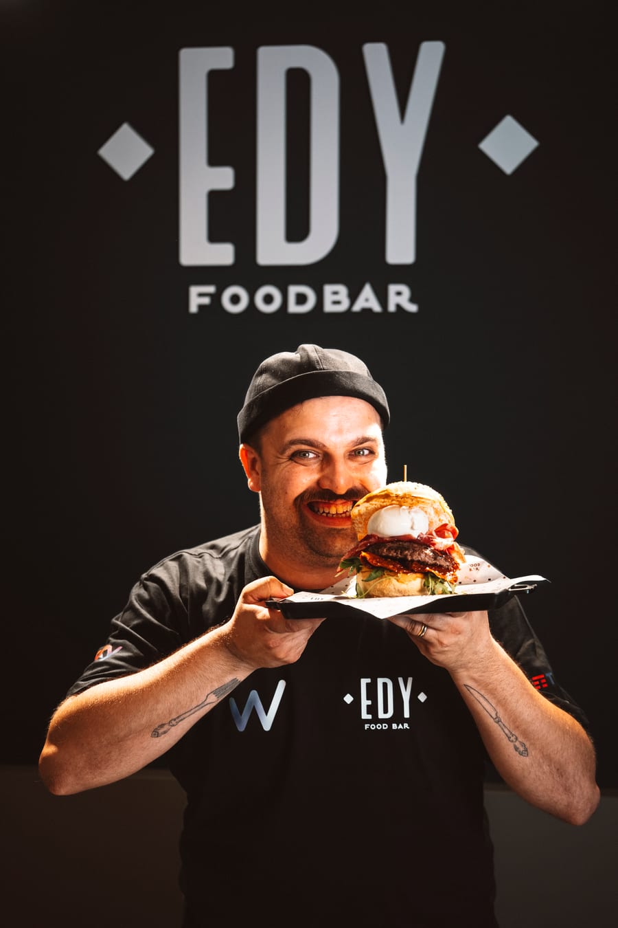 Il titolare sorride reggendo il vassoio col panino, sotto l’insegna EDY FOODBAR dipinta sulla parete nera