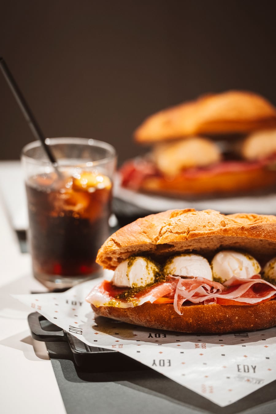 Il panino col crudo, la burrata e il pesto in primo piano, il bicchiere e un secondo panino sfocati dietro