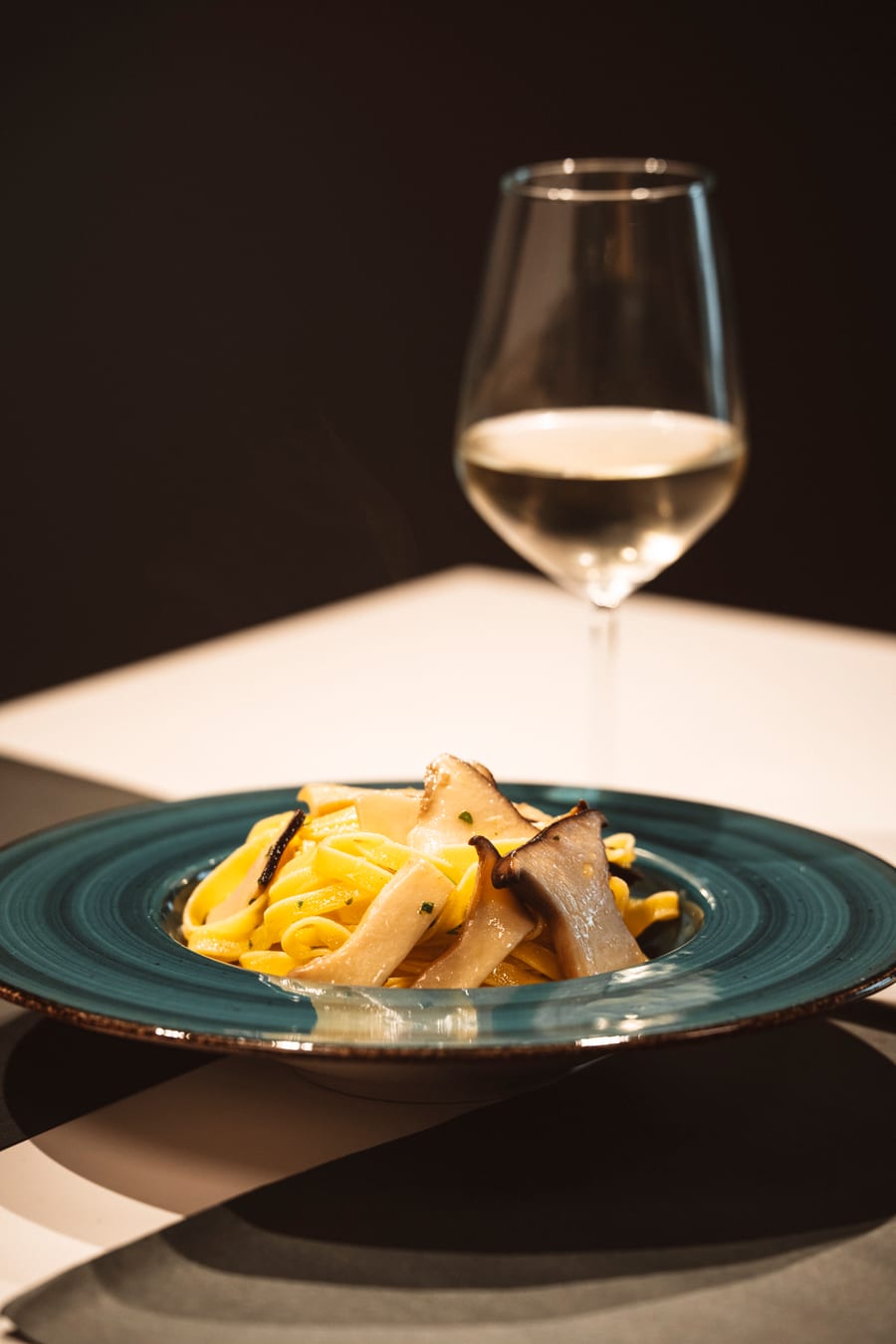 Le tagliatelle ai funghi cardoncelli nel piatto turchese, il calice di bianco dietro in controluce