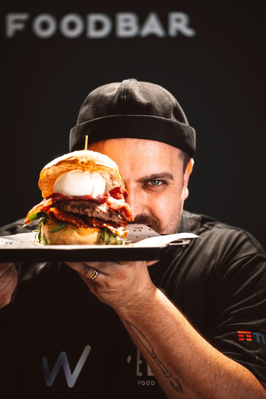 Il titolare regge il vassoio col panino davanti al viso, un occhio solo scoperto, sotto la scritta FOODBAR sulla parete nera