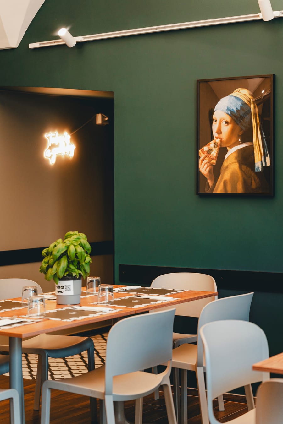 La sala dalla porta: la parete verde con la Ragazza con l’orecchino di perla che morde una pizza, un neon acceso e i tavoli apparecchiati col basilico