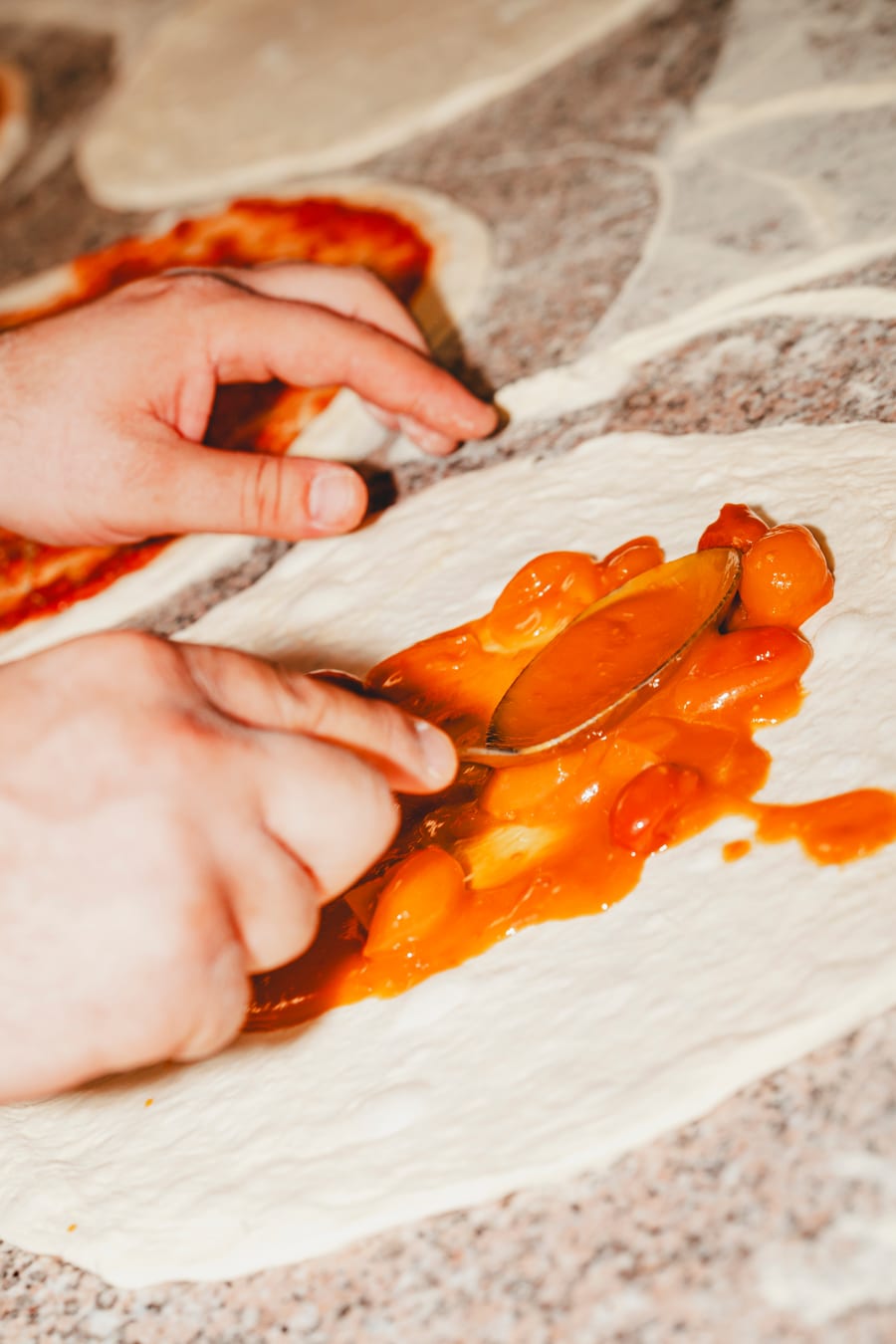 Due mani stendono con il cucchiaio il sugo di pomodorini sul disco di pasta, sul banco di granito
