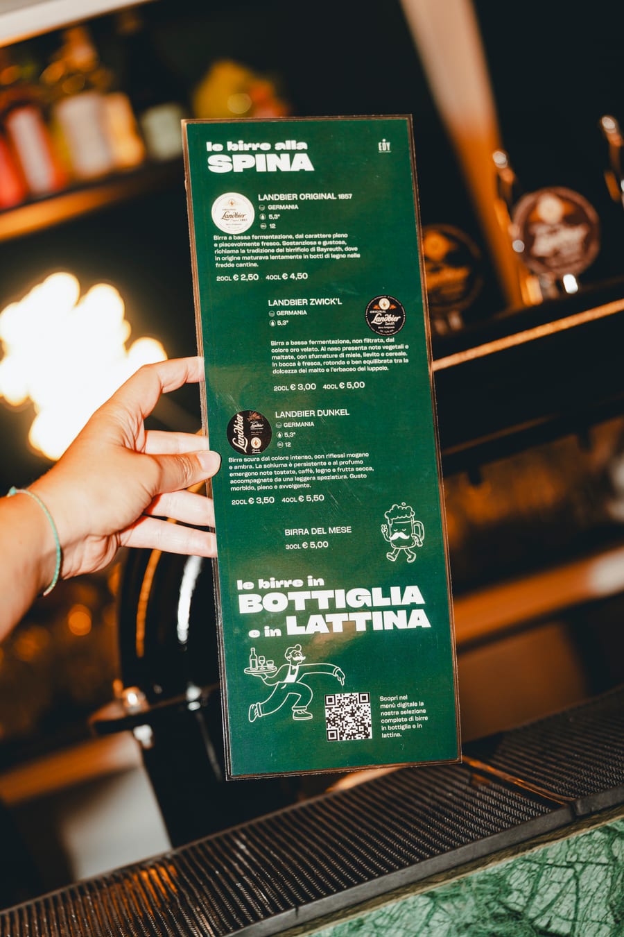 Una mano mostra al banco la lista verde delle birre alla spina e in bottiglia, illustrata col boccale coi baffi