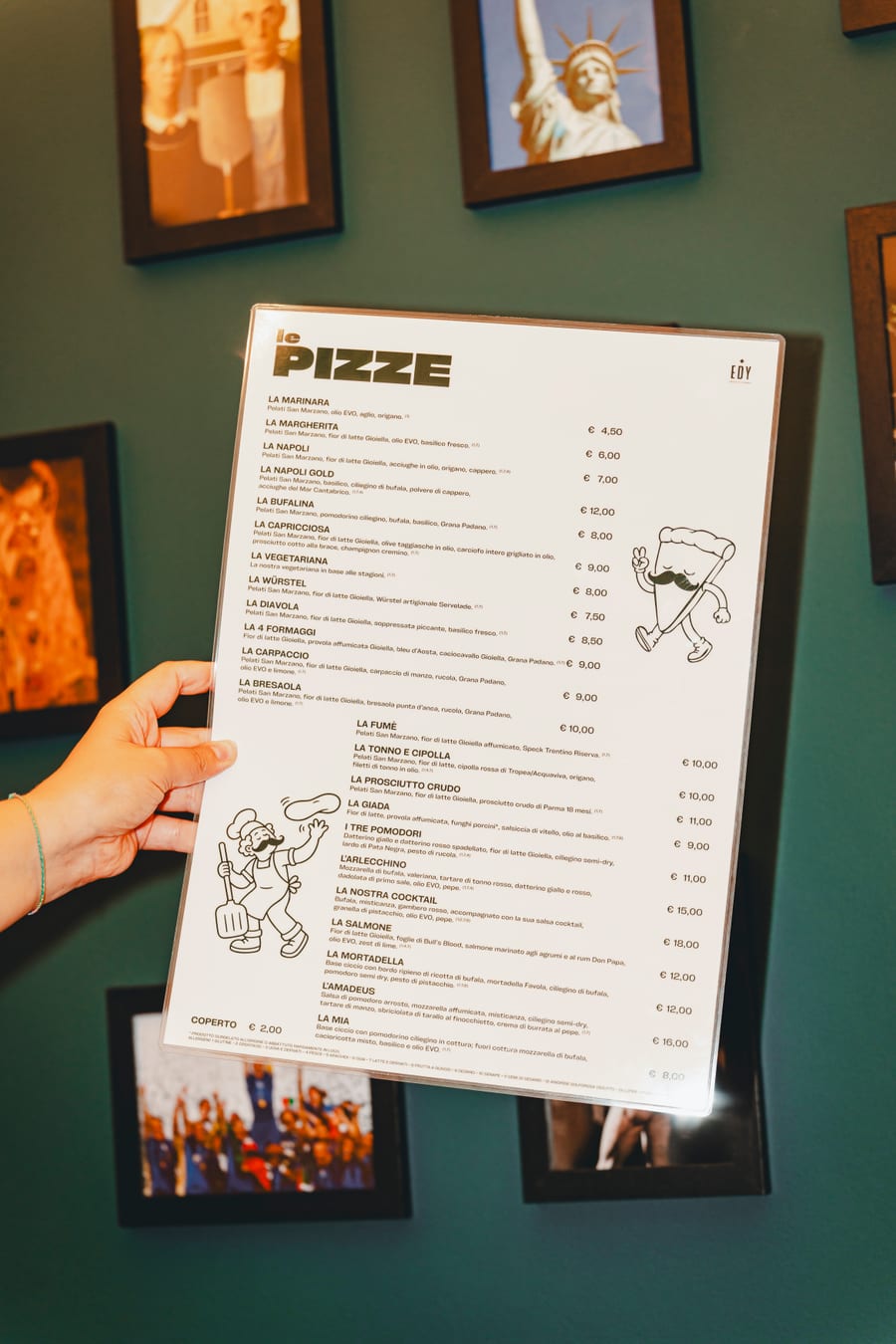 Il menù delle pizze tenuto in mano davanti alla parete verde con le cornici, con la fetta coi baffi disegnata a margine