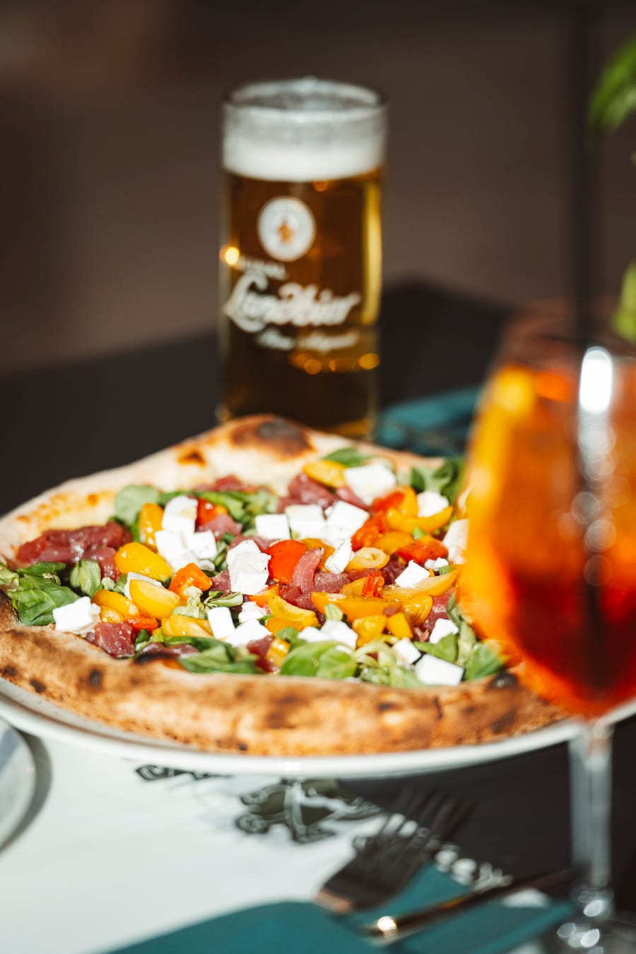 La pizza col songino, i pomodorini gialli e i dadi di formaggio sul tavolo, fra un boccale di birra e uno spritz