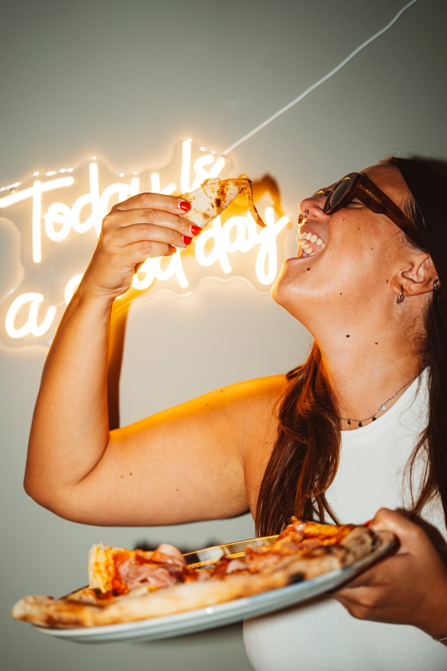 Una ragazza con gli occhiali da sole si cala in bocca una fetta di pizza davanti al neon «Today is a good day»
