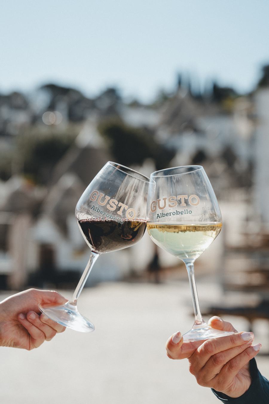 Due calici marchiati Gusto Alberobello, rosso e bianco, brindano davanti ai trulli
