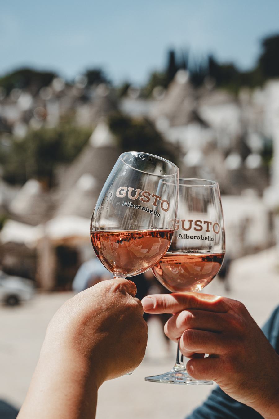 Brindisi con due calici di rosato marchiati Gusto Alberobello, i trulli sullo sfondo