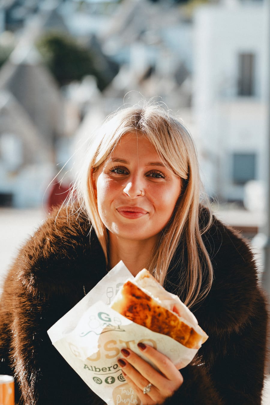Una ragazza sorride con una focaccia avvolta nella carta di Gusto, fra i trulli