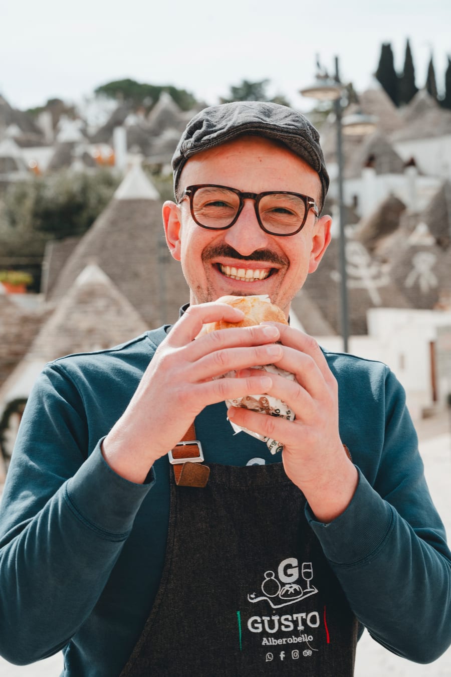 Pasqualino, col grembiule di Gusto, sorride con un panino fra le mani davanti ai trulli