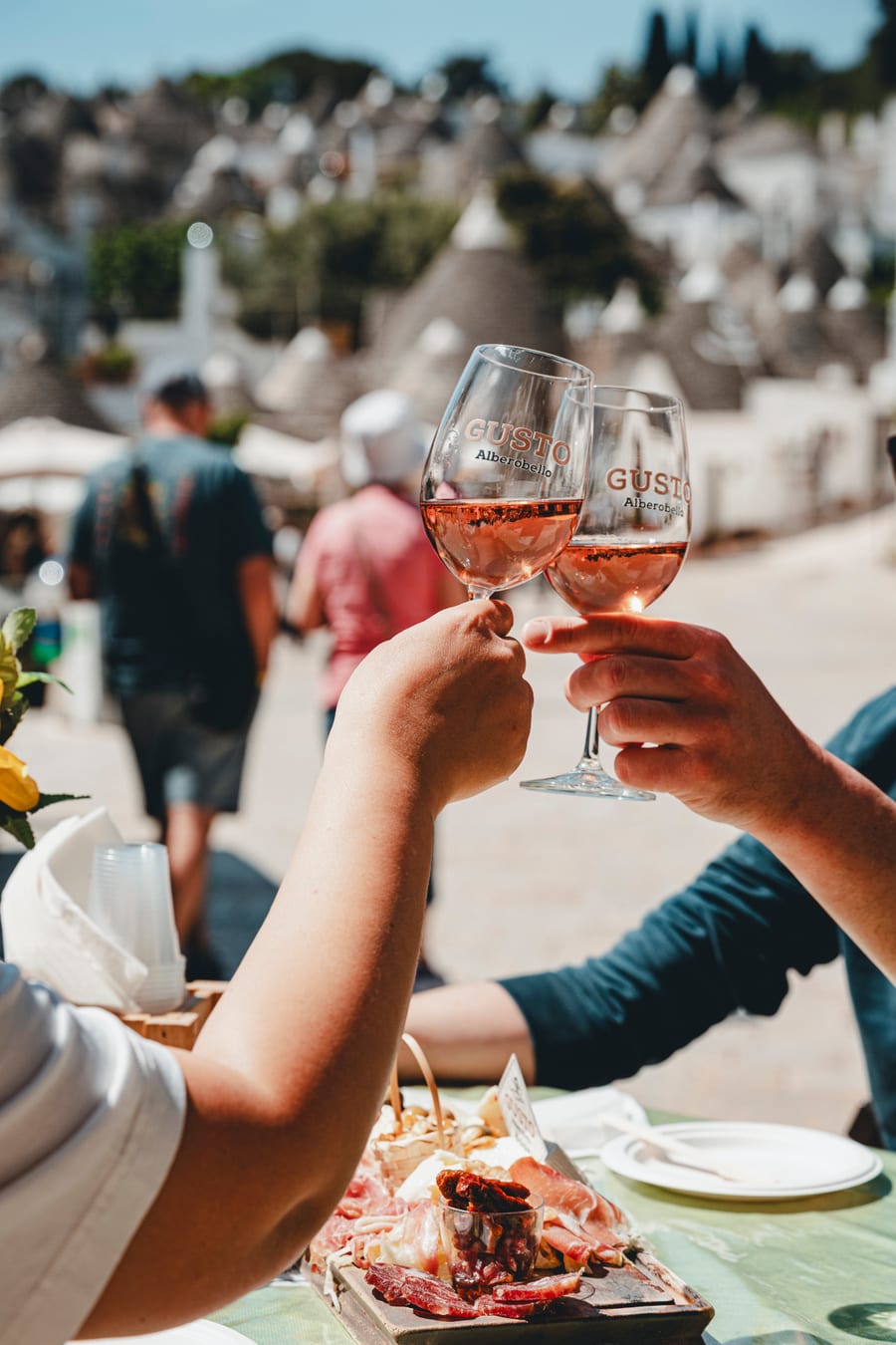Due calici di rosato brindano sopra il tavolo, fra la gente che passeggia ad Alberobello