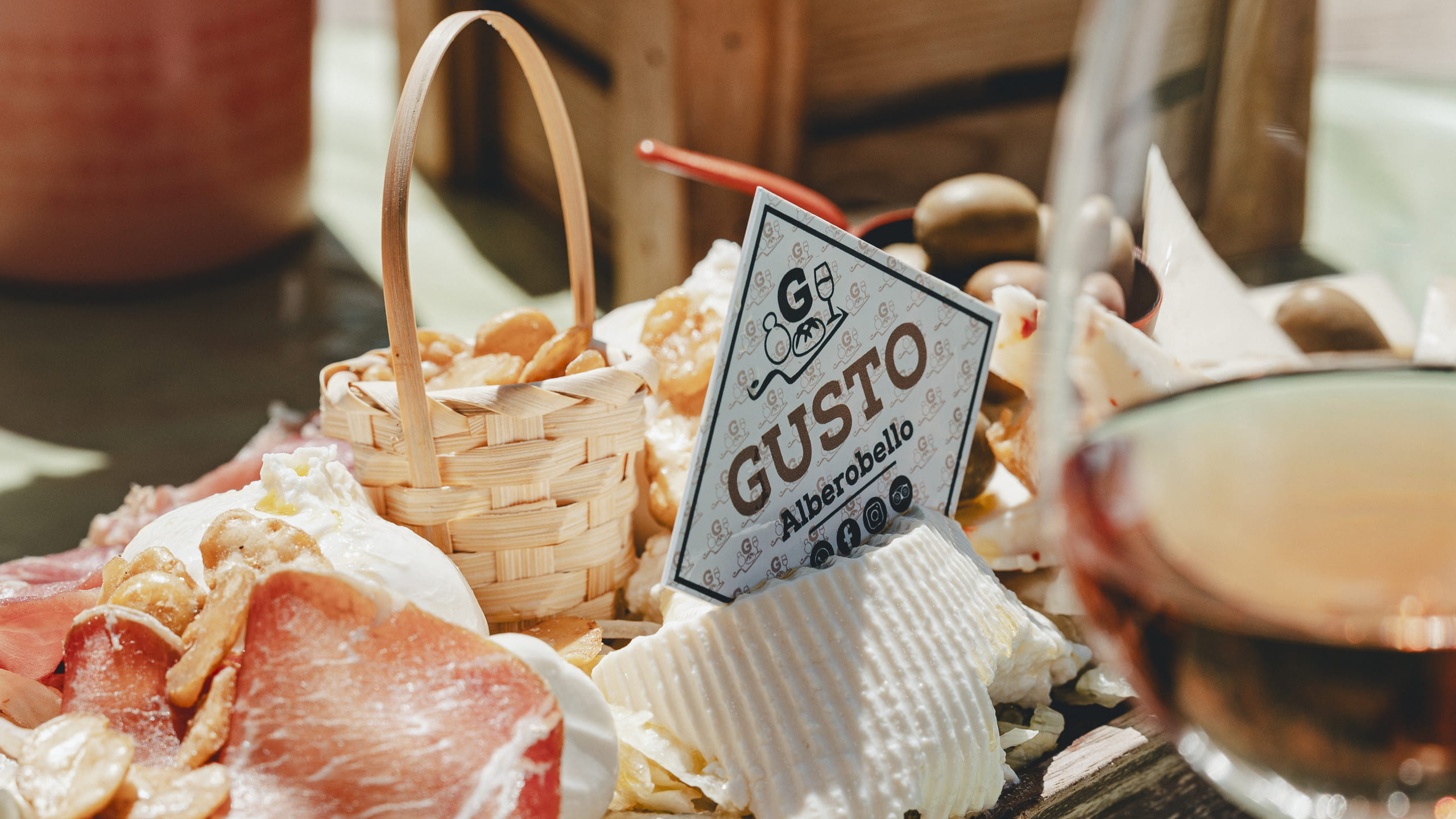 Tagliere di salumi, burrata e taralli con il cartoncino Gusto Alberobello, accanto a un calice di rosato