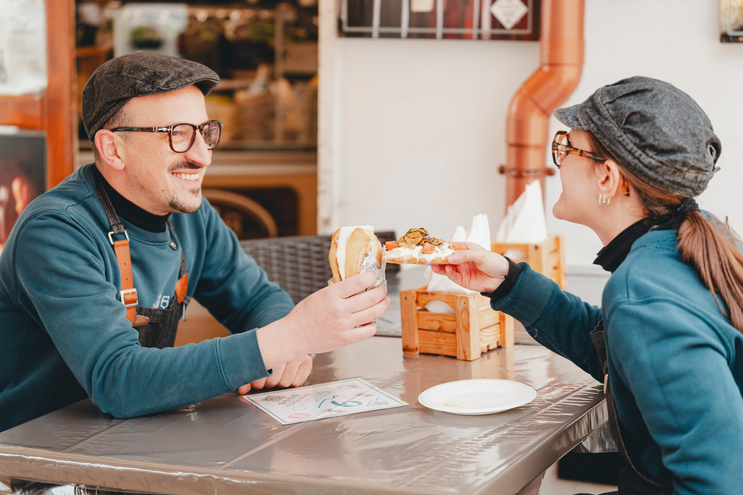 Due soci di Gusto si passano una focaccia al tavolino davanti al locale