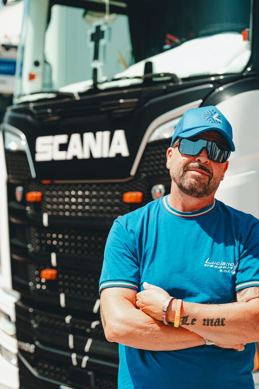 Un autista con cappellino e occhiali da sole, a braccia conserte davanti al muso nero di uno Scania