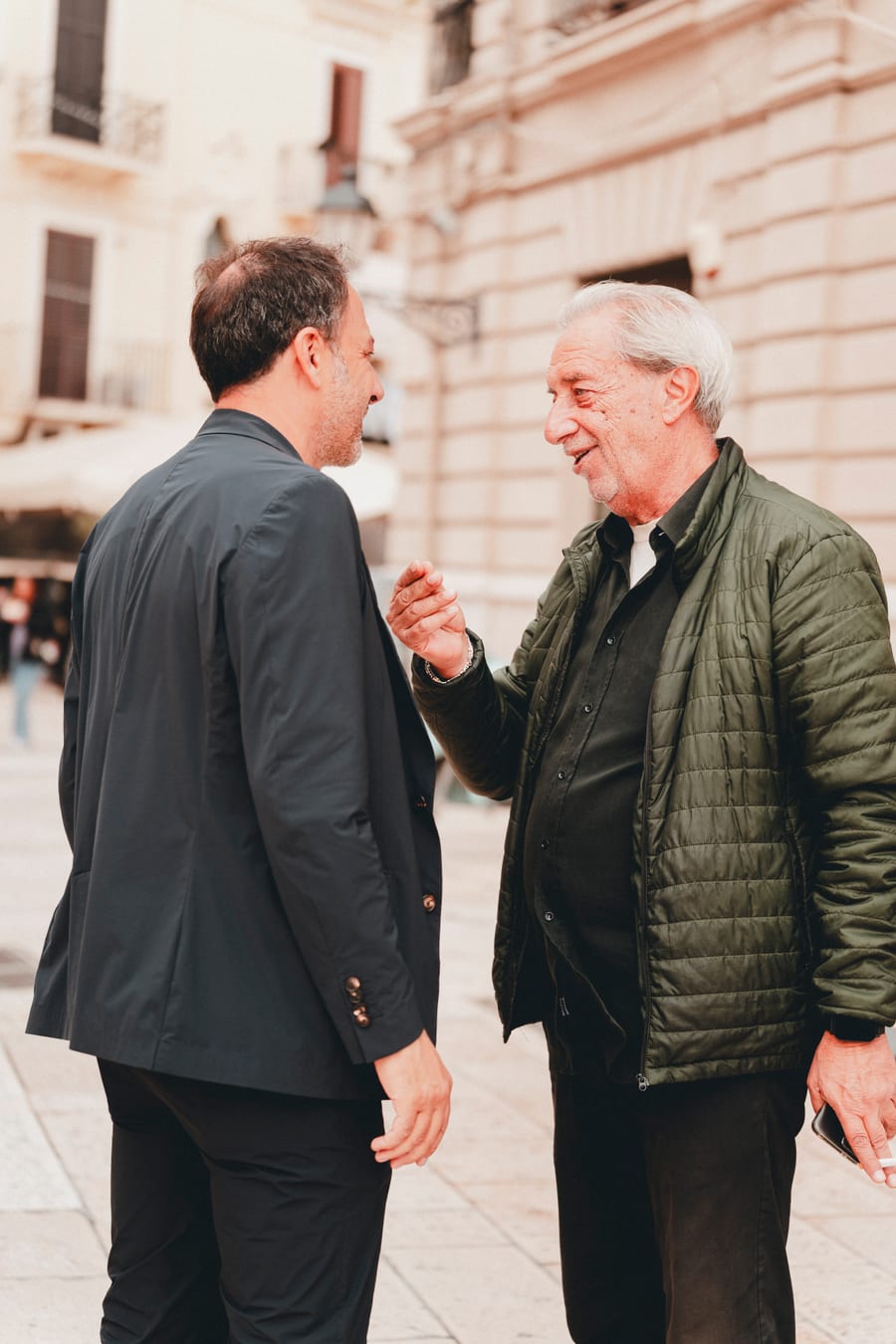 Di spalle in piazza, mentre un signore anziano in giacca verde gli parla indicandolo