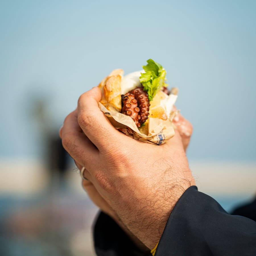 Due mani stringono il panino col polpo nella carta, contro il mare sfocato