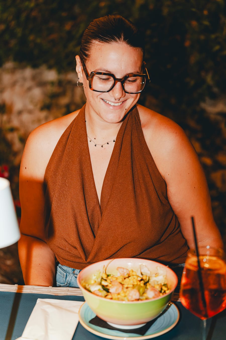 Una ragazza sorride al tavolo la sera, davanti a una ciotola e allo spritz