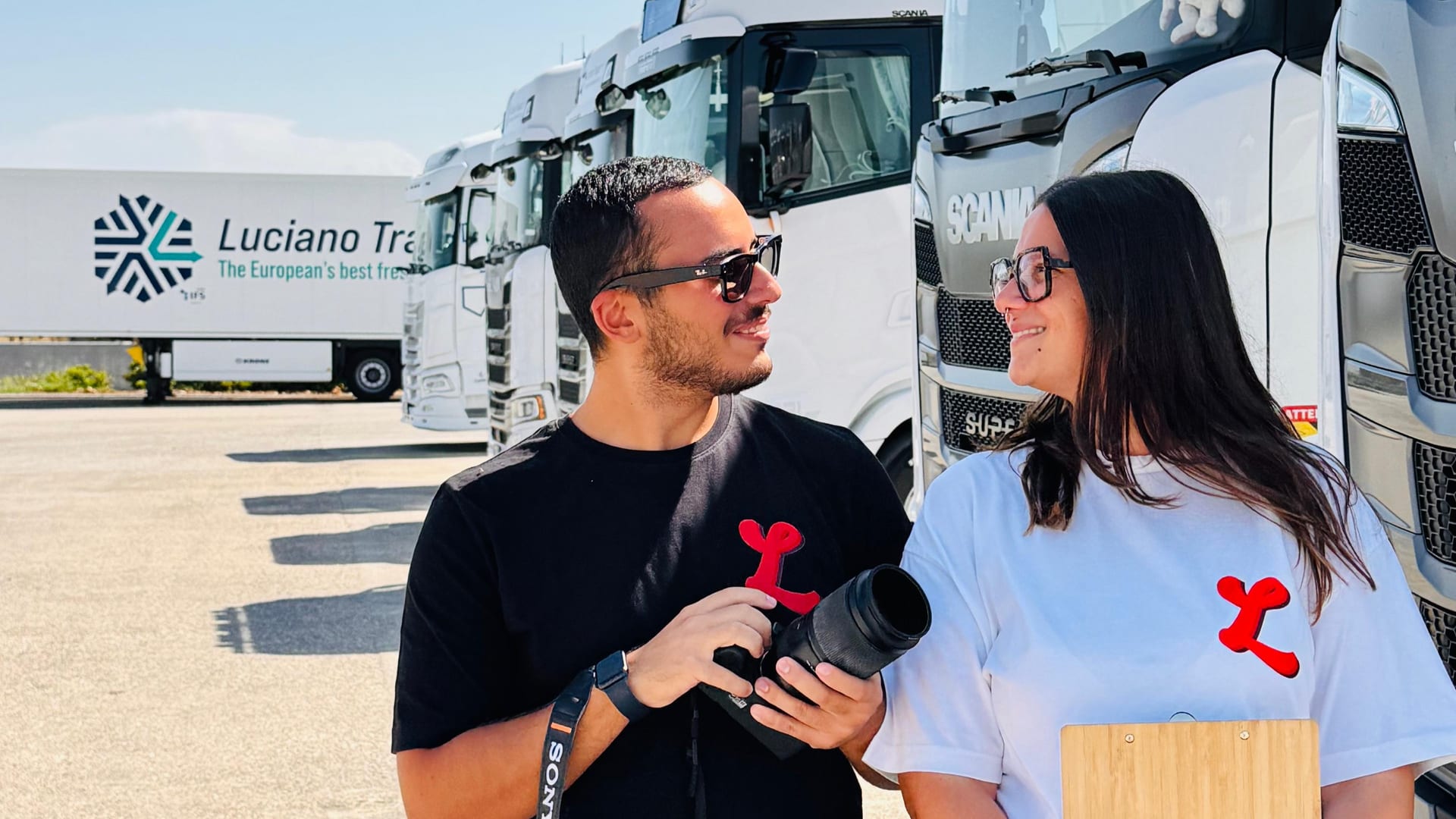 Alessandro Lupelli e Asia Cafueri che si sorridono davanti ai camion di Luciano Trasporti, la L rossa dello studio sulle magliette: lui con la macchina fotografica, lei con una cartellina